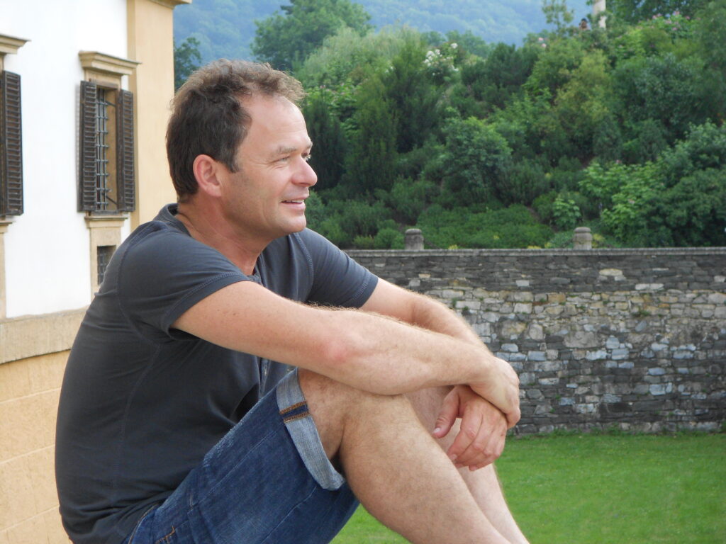 Foto von Markus Hirtler sitzent im Garten im Hintergrund ein Haus ein Steinmauer und viele Pflanzen