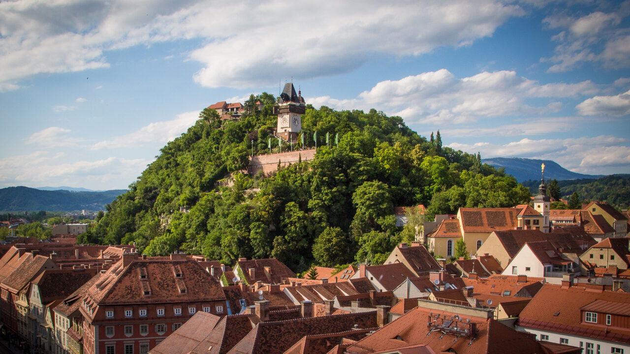Ein Foto des Grazer Schlossberg im Zentrum, im unteren Bereich die Dächer der Grazer Altstadt. Blauer Himmel mit weißen Wolken.