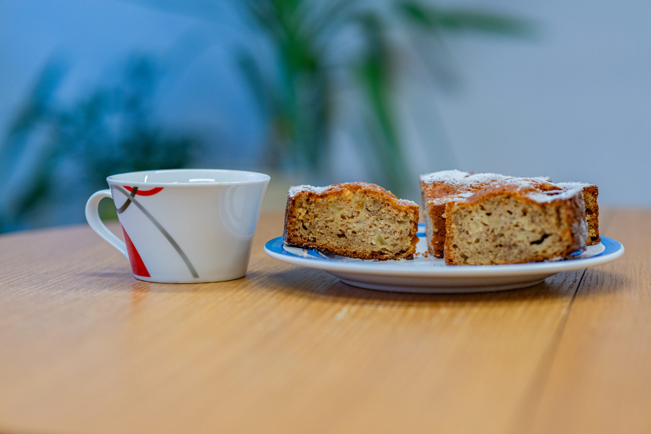 Auf dem Bild befindet sich eine Tasse. Rechts daneben steht ein Teller mit Kuchen auf einem Holztisch.