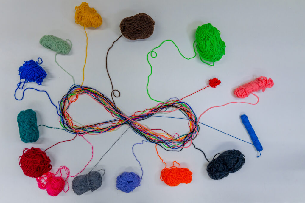 Eine bunte, liegende Acht wird aus verschiedenfarbigen, von außen zulaufenden Fäden gebildet.