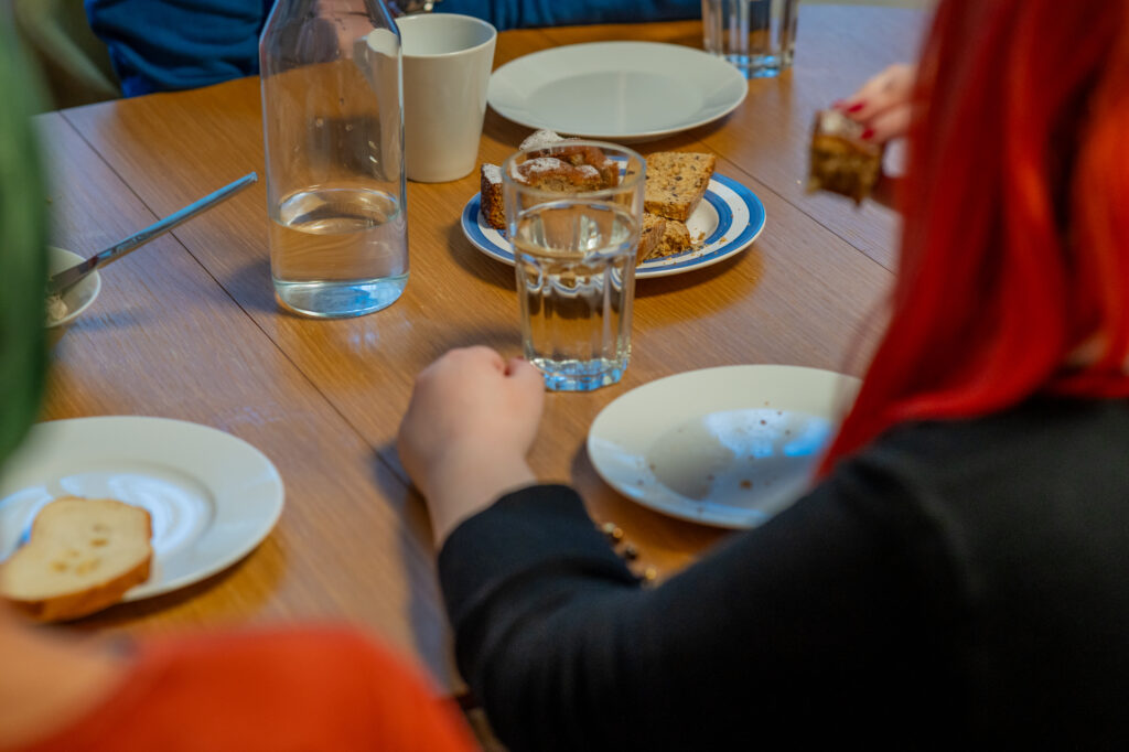 Um einen Tisch sitzen Personen, die sich unterhalten. Am Tisch stehen Teller und Gläser. In der Mitte befindet sich ein Teller mit Kuchenstücken.
